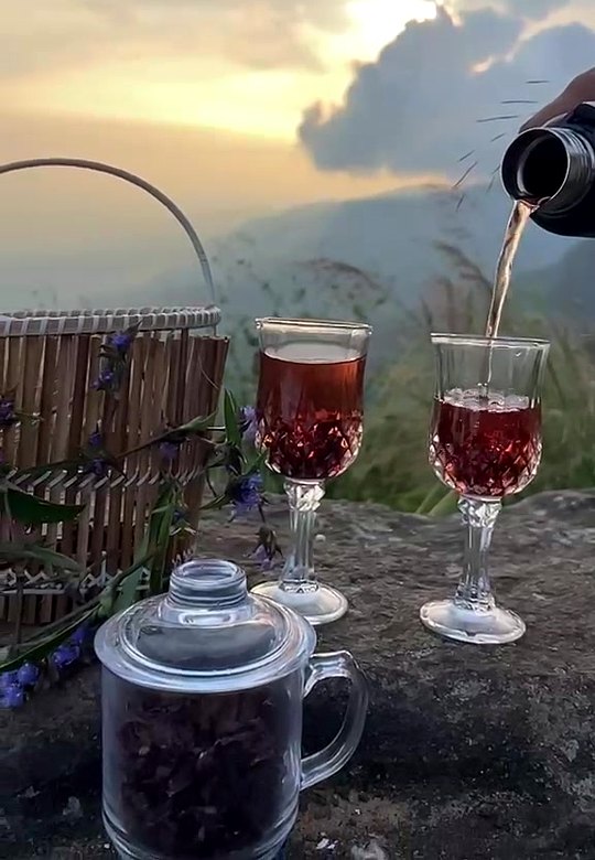 Roselle tea being poured into glasses beside a jar of dried calyces and a bamboo basket, at sunset